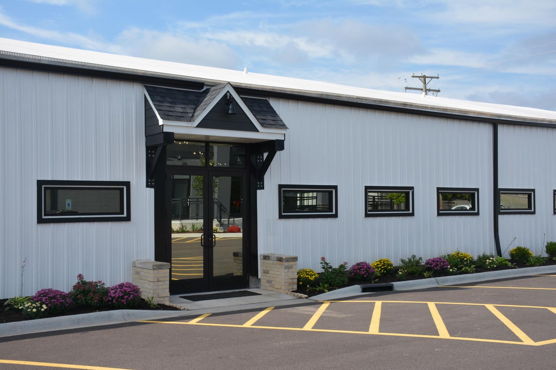 White commercial building with a black-trimmed entryway, horizontal windows, and flower beds in a parking lot.