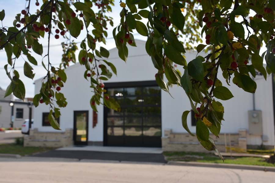 A white warehouse-style building with a black glass garage door and stone accents, framed by leafy green tree branches.