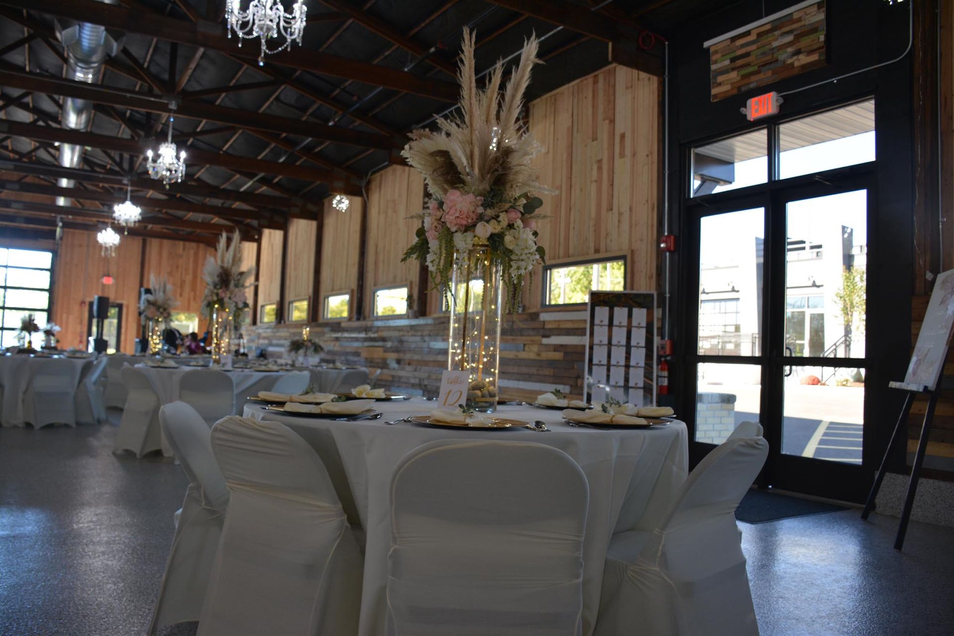 A wedding reception hall features round tables with white tablecloths, centerpieces of dried flowers, and crystal lights.