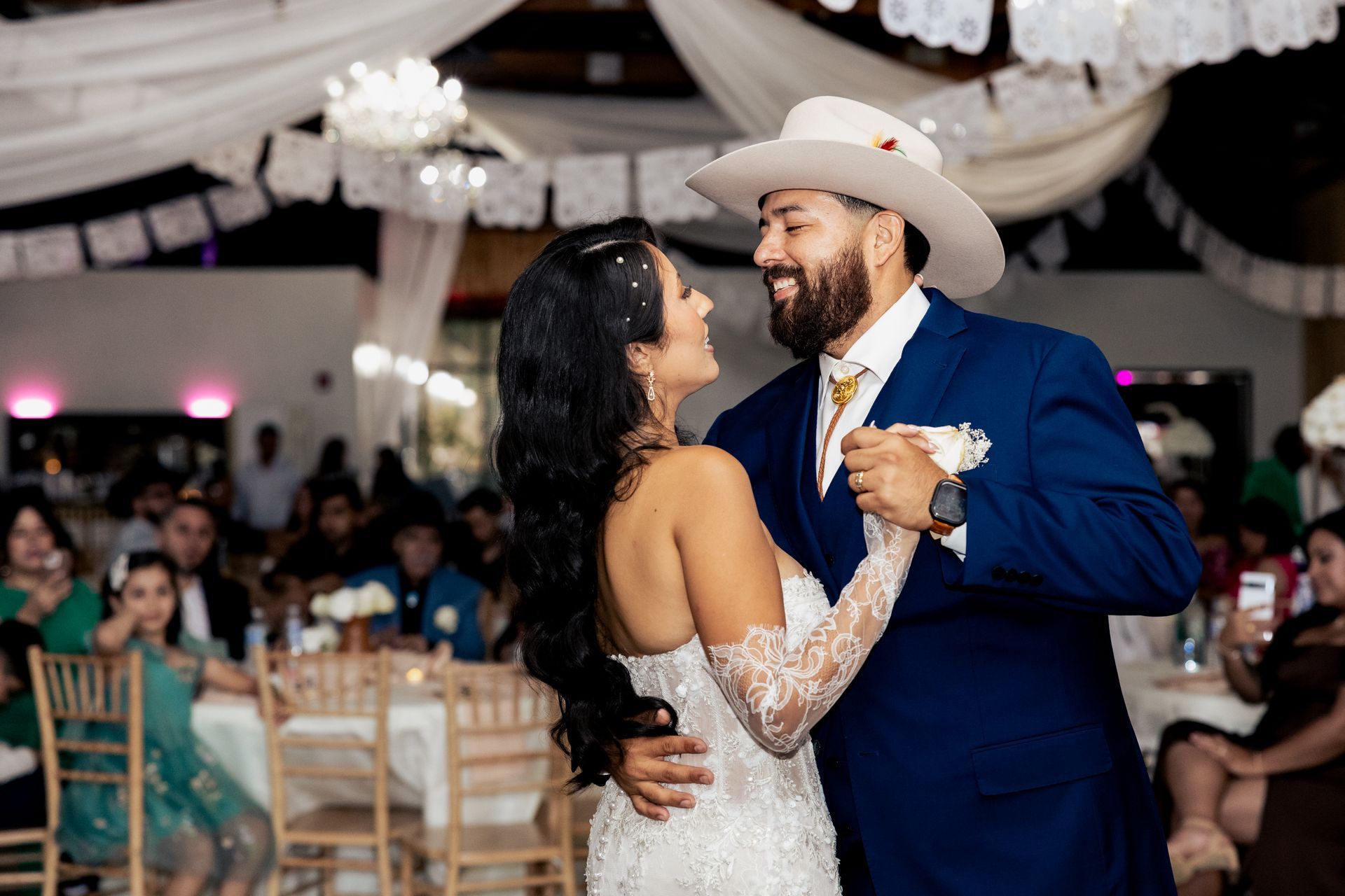 A bride in a lace gown and groom in a blue suit and cowboy hat share a dance at a wedding reception.