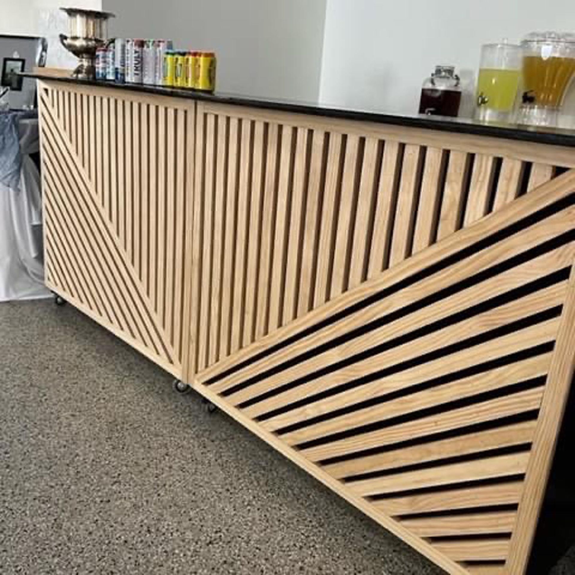 A modern, portable wooden bar with geometric, diagonal slat detailing sitting on a speckled concrete floor.