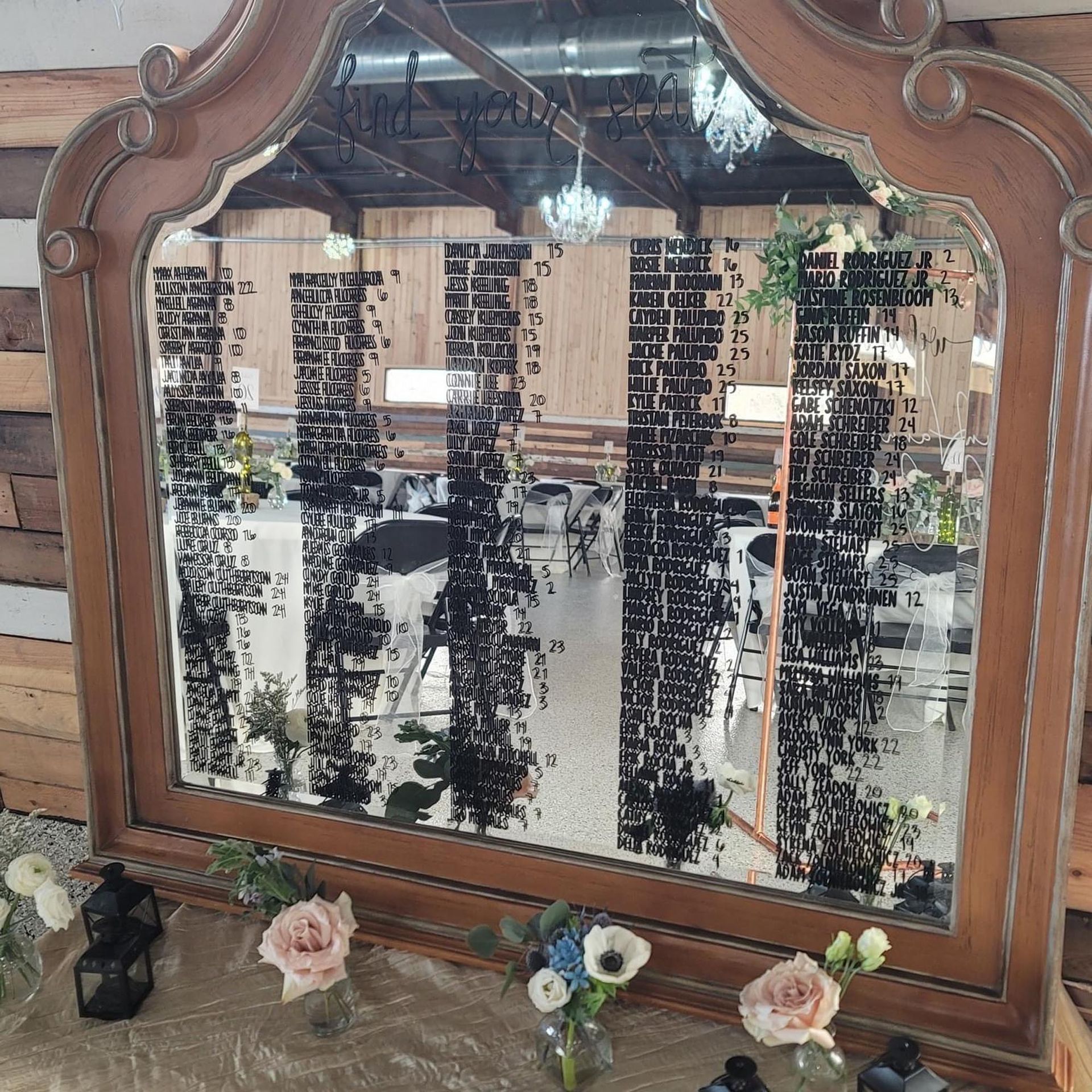A large, ornate wooden mirror displays a guest seating chart with black text, set on a table decorated with flowers.