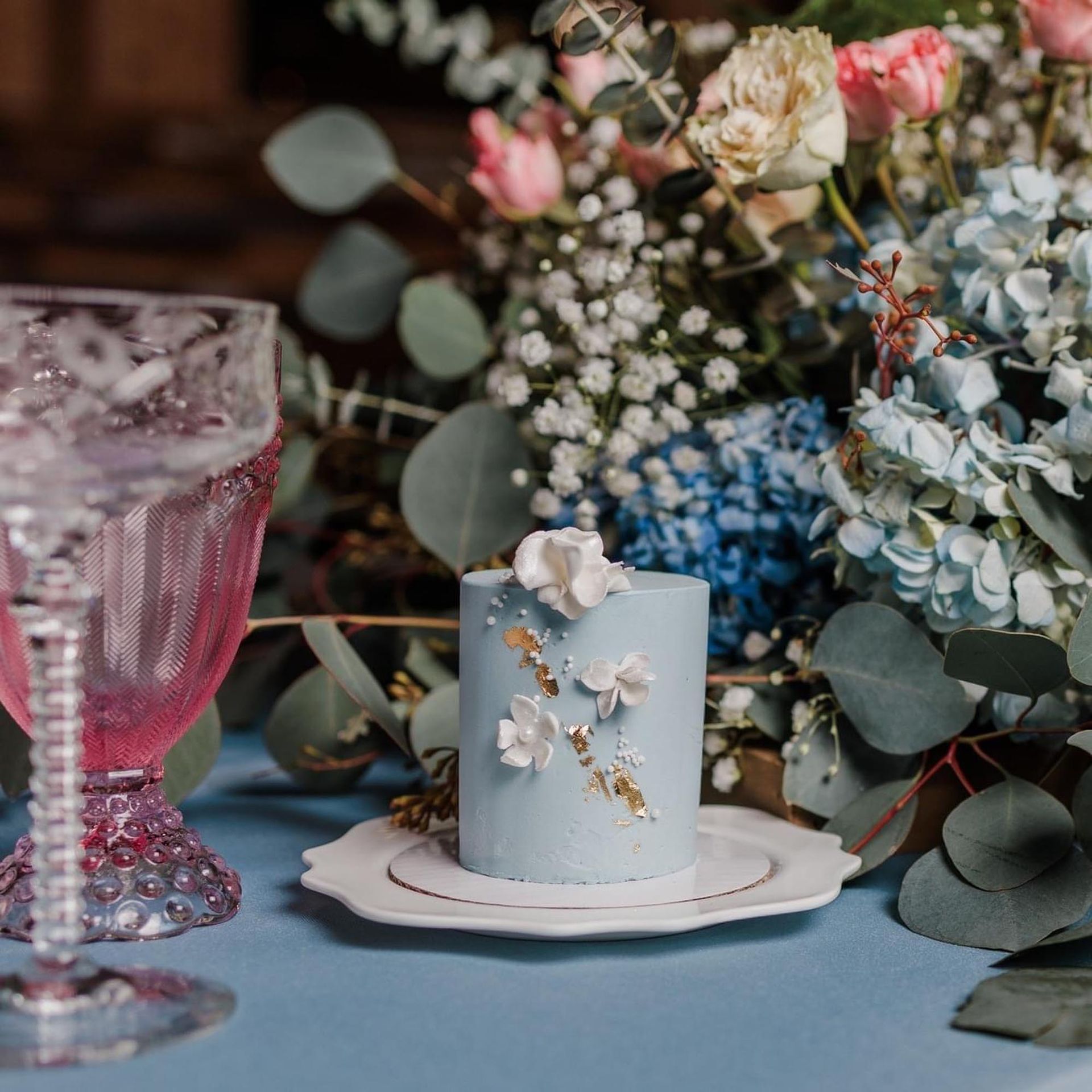 A small, light blue cake decorated with white flowers and gold leaf sits on a plate before a bouquet of flowers.