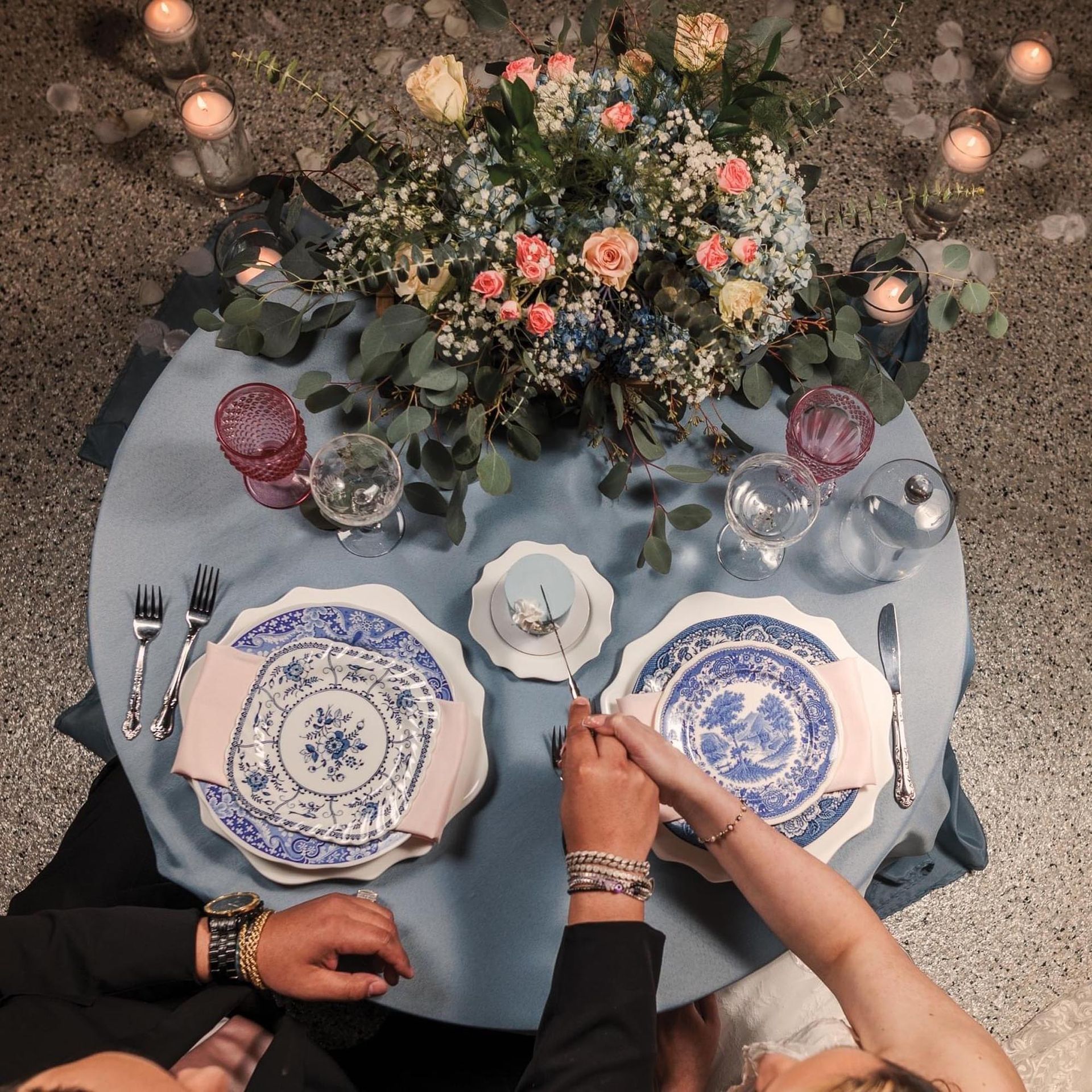 A couple holds hands over a blue-clothed table set for two with floral arrangements, candles, and vintage patterned plates.