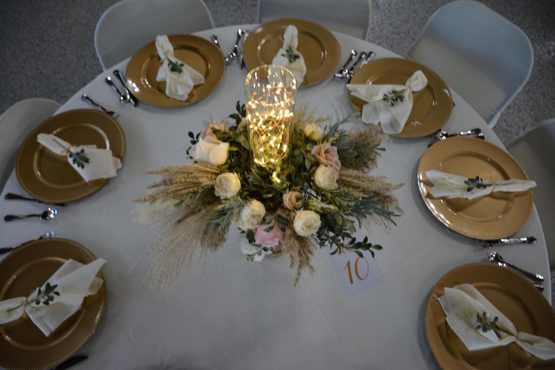 A round table is set with gold charger plates, white napkins with greenery, and a floral centerpiece with fairy lights.