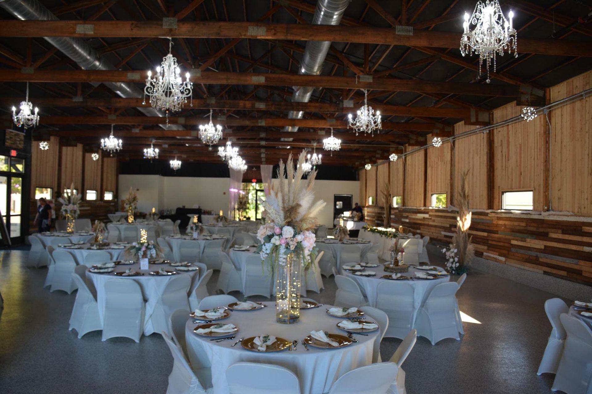 A banquet hall with round tables covered in white linens, decorated with floral centerpieces under hanging chandeliers.