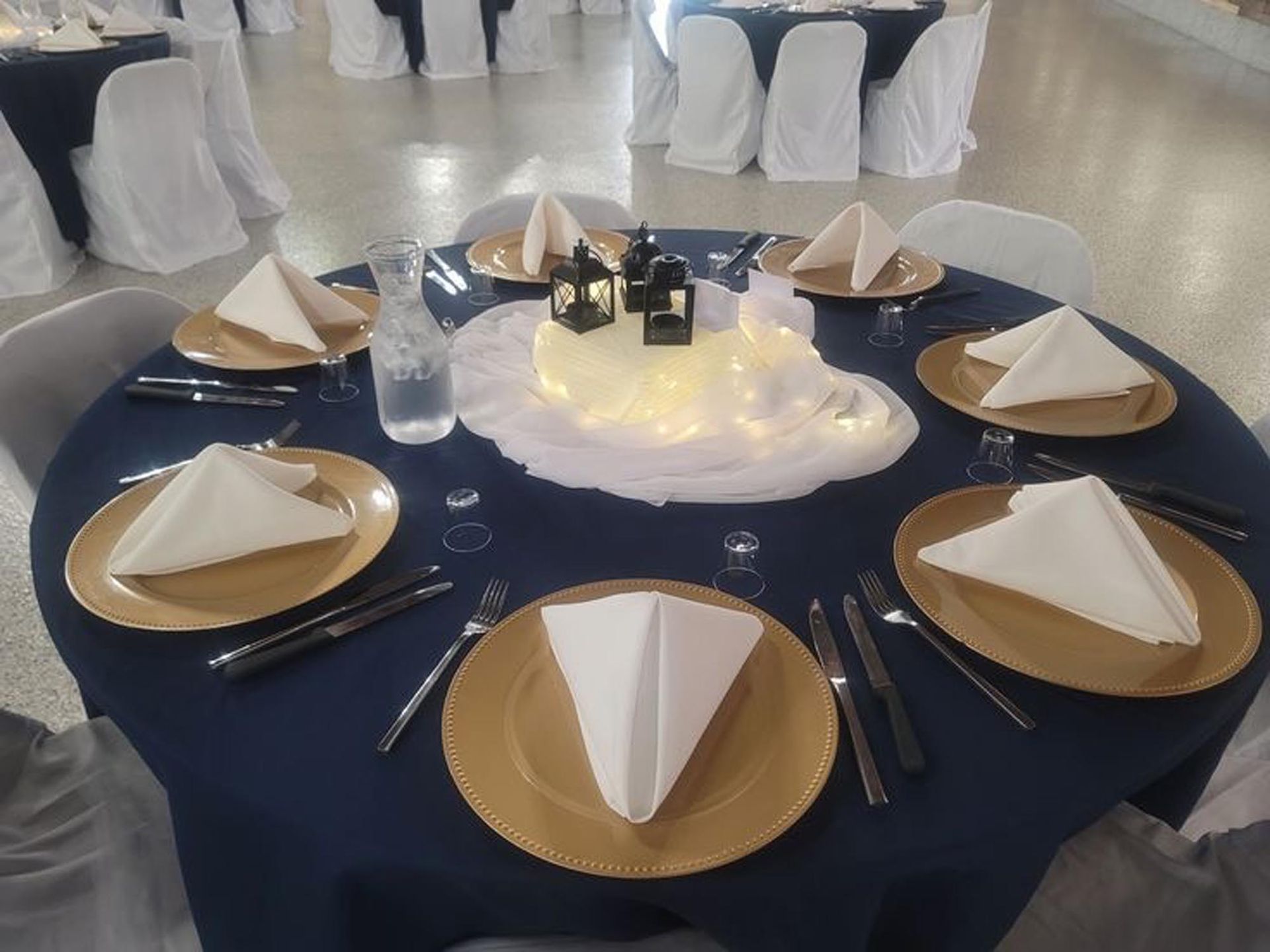 A round table set for a formal event with a navy tablecloth, gold chargers, white napkins, and a center floral arrangement.