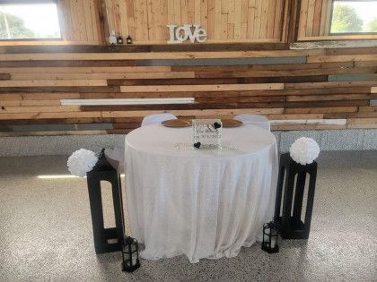 A white-covered round sweetheart table is set in front of a wood-paneled wall, flanked by two black lantern stands.