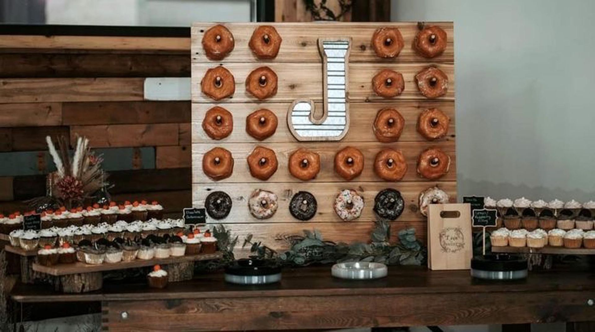 A wooden table displays a wall of donuts with a metal letter J, flanked by tiered stands filled with small desserts.