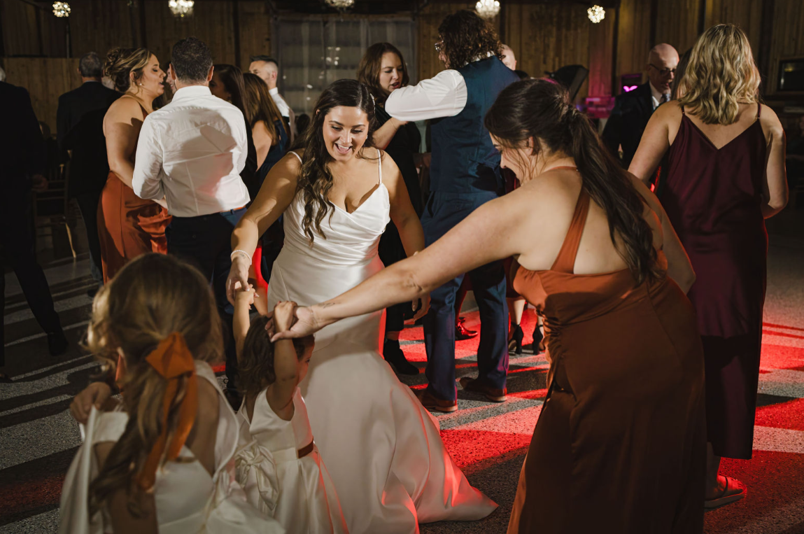 People in formal attire dance joyfully on a lit-up dance floor in a rustic indoor wedding reception setting.