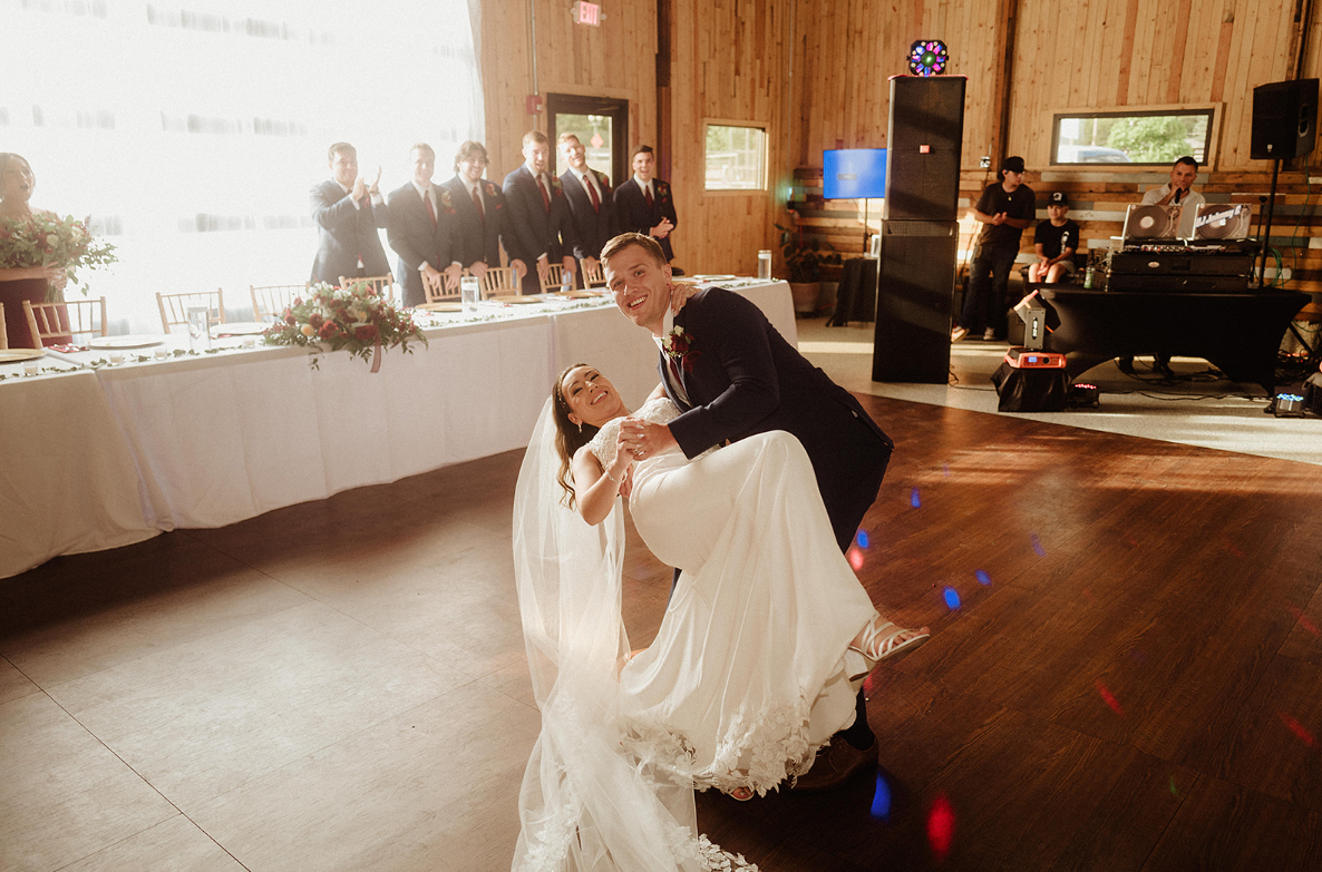 A groom dips his bride in a white wedding dress during their first dance on a wooden floor in a rustic barn venue.