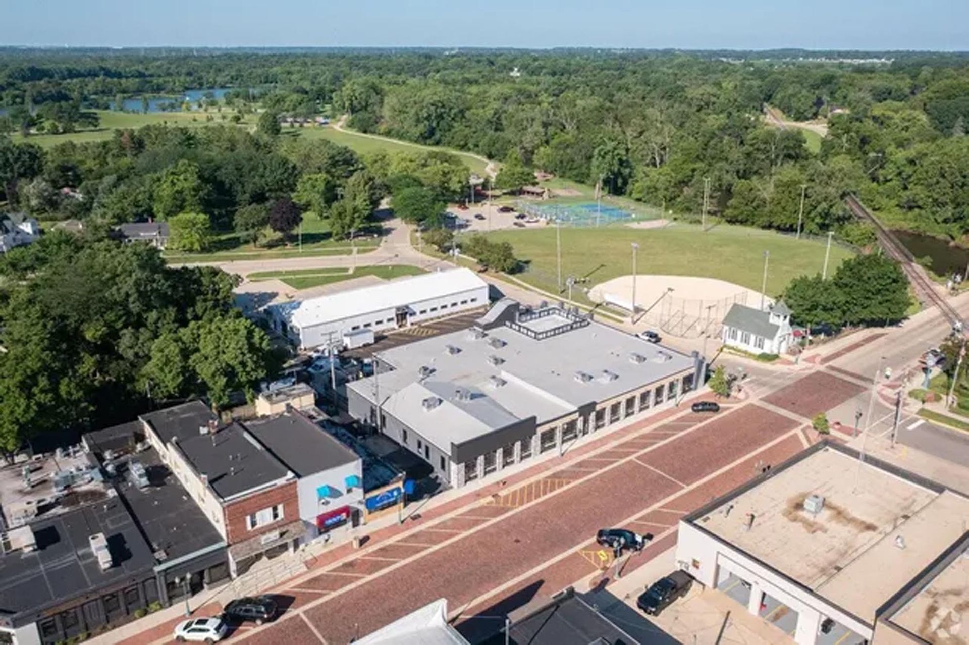 Aerial view of a small town street, brick-paved road, multi-story buildings, and a park with a baseball diamond.