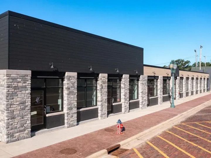 A modern single-story commercial building with black siding and stone pillars, located along a brick sidewalk and parking.