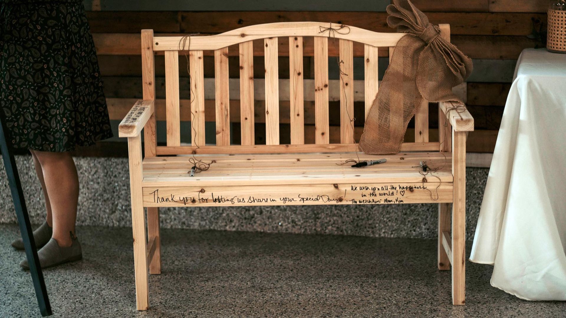 A wooden guestbook bench with signatures written on the seat, placed indoors next to a table with a white cloth.