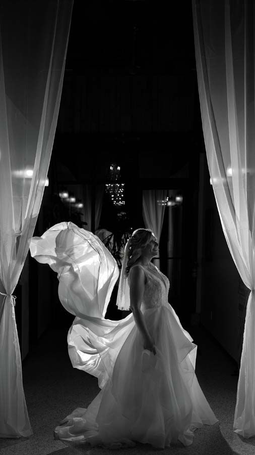 A bride stands between draped curtains, the train of her gown billowing out to her side in a dramatic black and white shot.