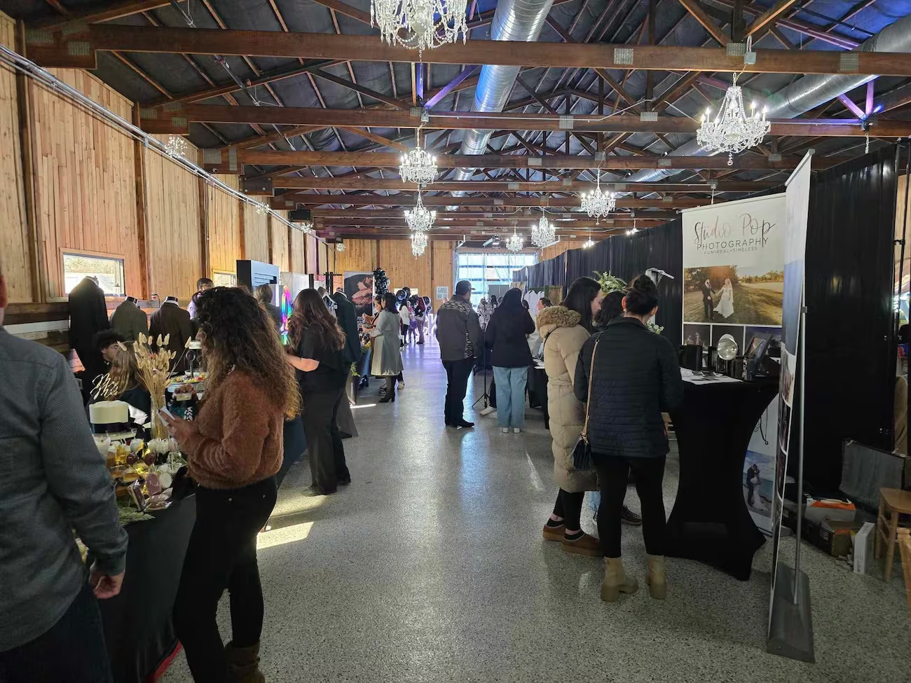 People browse vendor stalls at an indoor wedding expo in a rustic venue with high ceilings and crystal chandeliers.