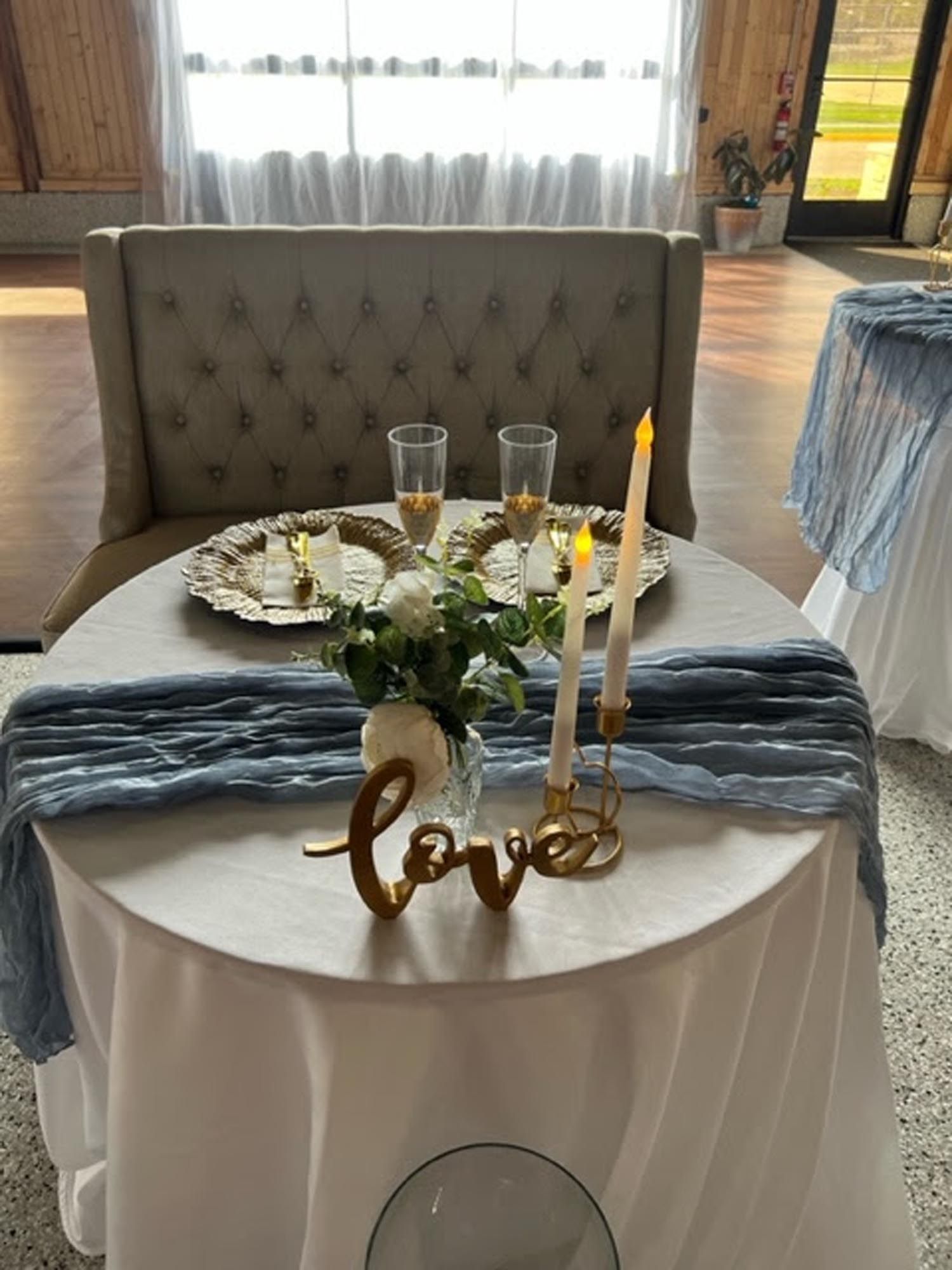 A small, round sweetheart table with white linens, a blue runner, candles, and a gold 