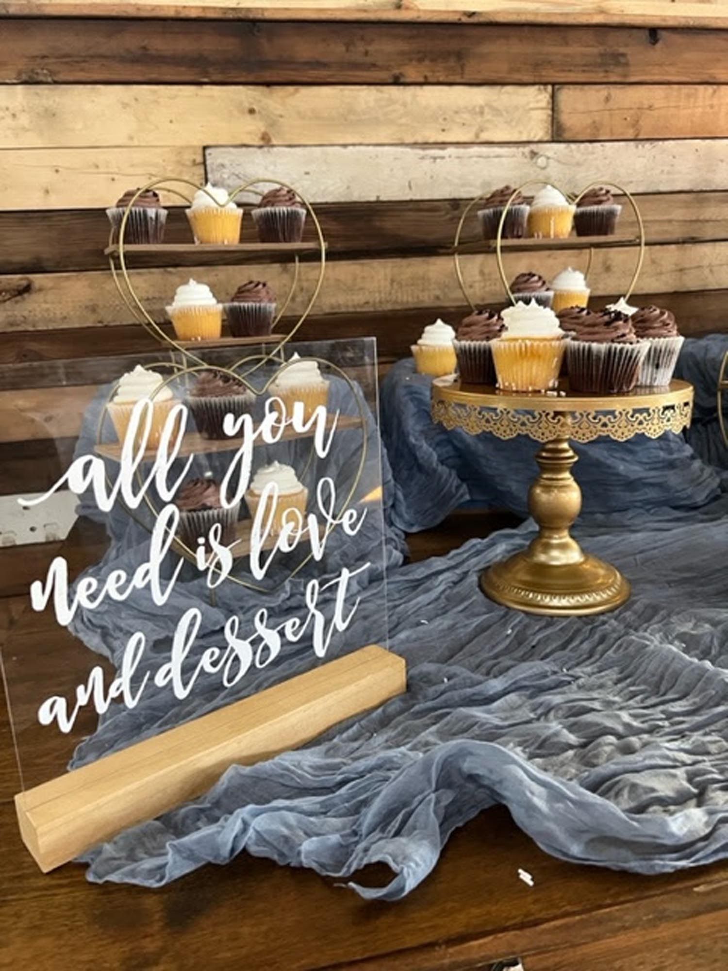 A dessert table featuring cupcakes on gold tiered stands and a clear sign that reads, 