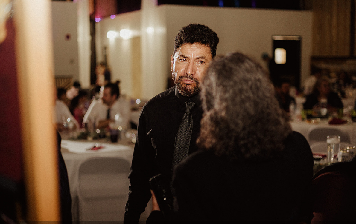 A person in a black shirt and tie talking to someone in a dark jacket in a dimly lit banquet hall with tables.
