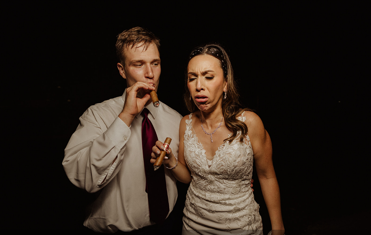 A person in a suit and a person in a white wedding dress hold cigars against a dark background, the latter grimacing.