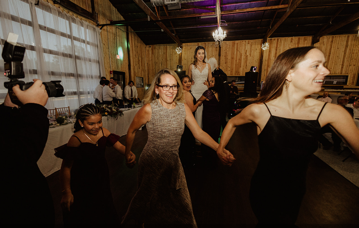 Three people hold hands and dance in a rustic event hall, with a wedding photographer capturing the moment on the left.