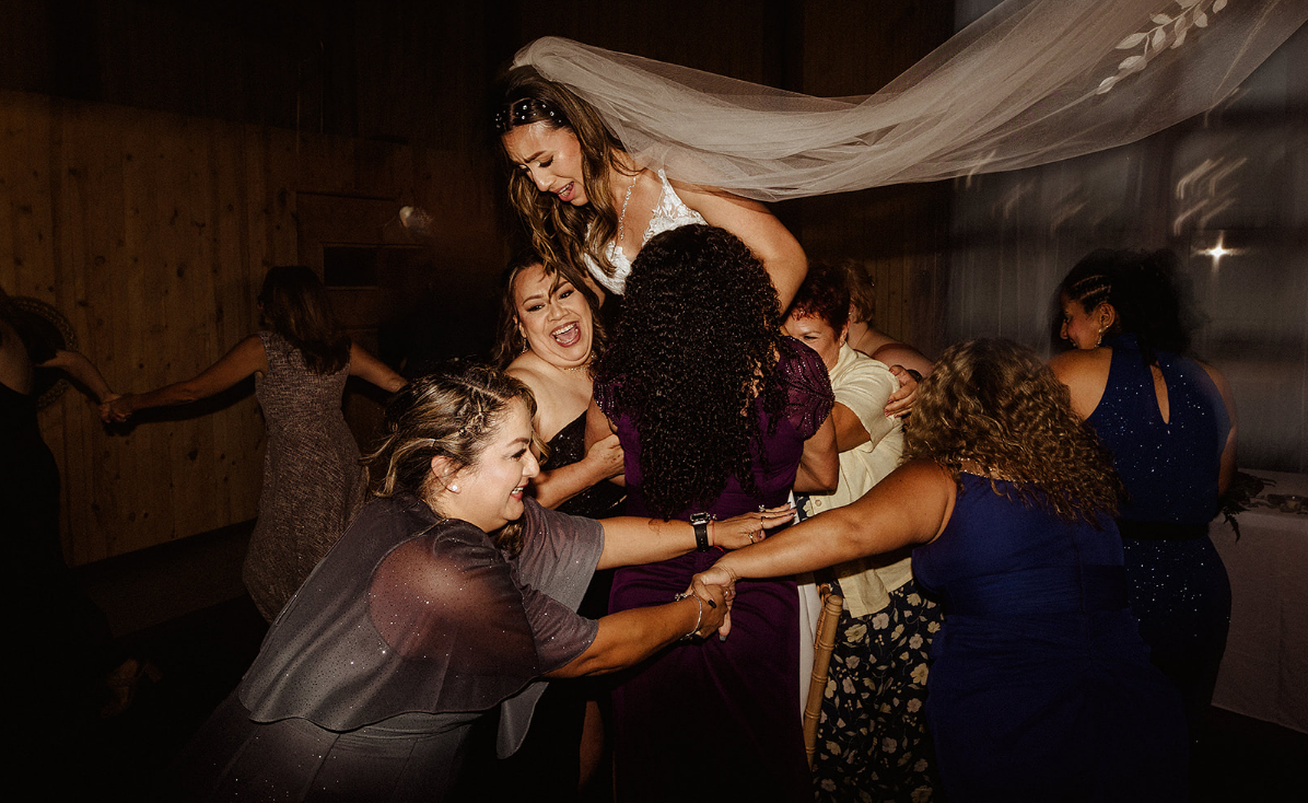 A bride is held aloft by a group of people at a dimly lit indoor celebration.