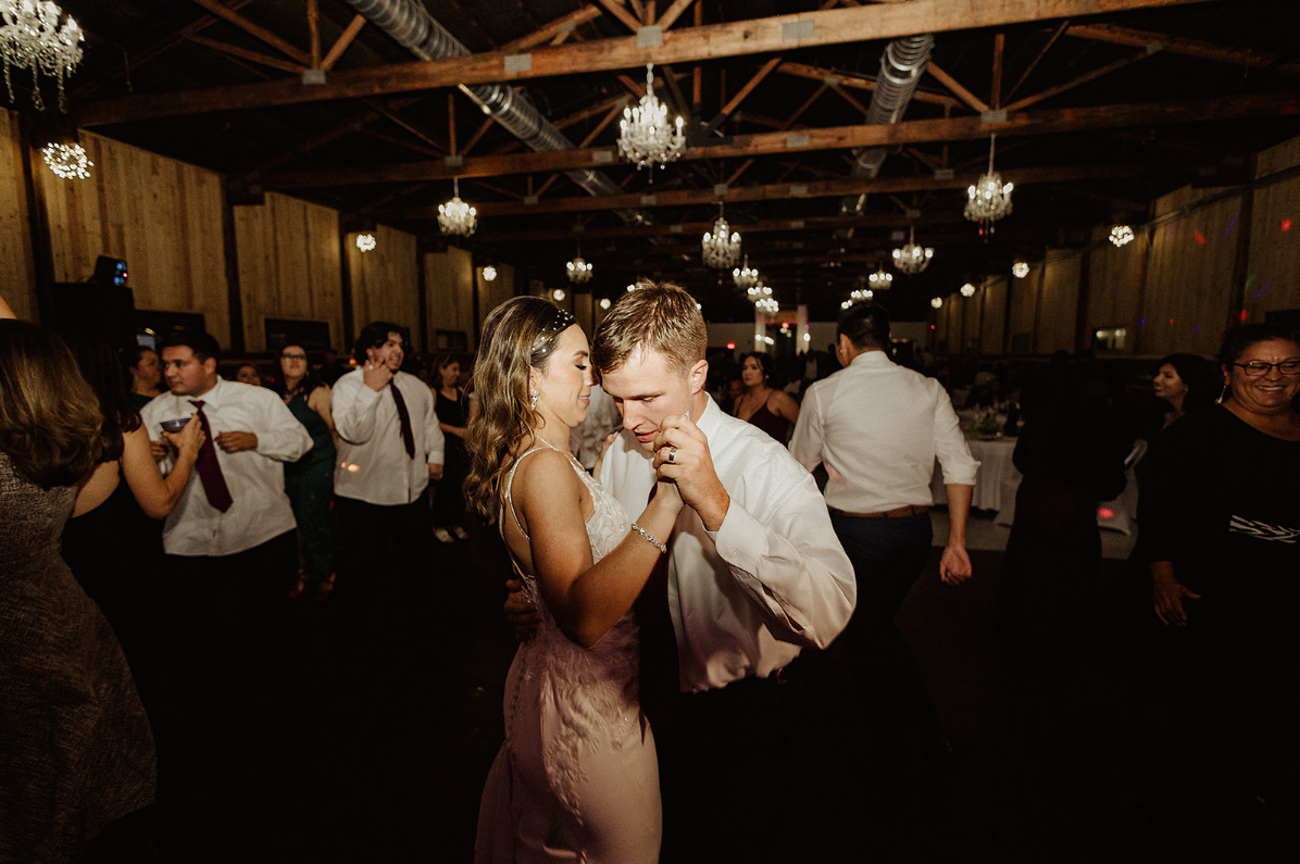 A couple dances together in a dimly lit rustic event hall filled with hanging chandeliers and wedding guests.