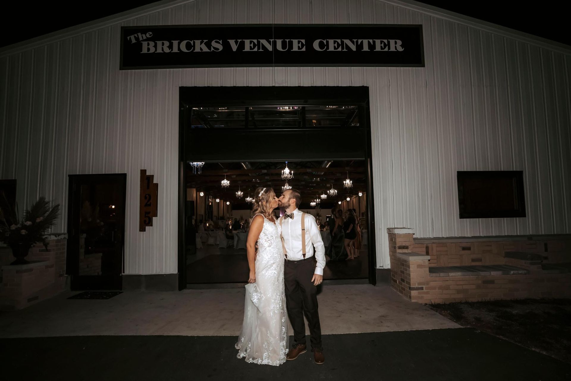 A couple kisses in front of the entrance to The Bricks Venue Center at night, illuminated by interior lights.