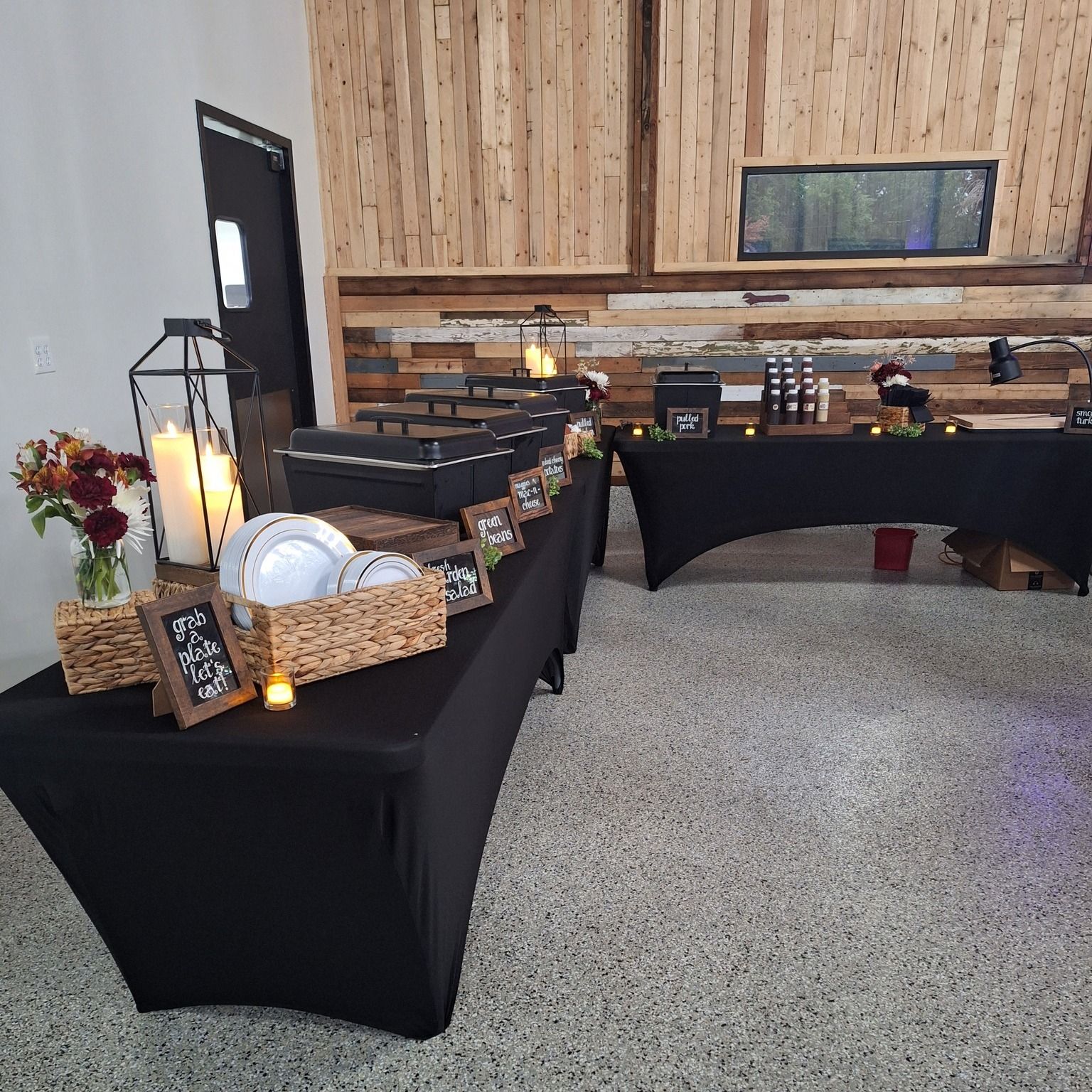 A buffet-style setup with black-covered tables, plates in woven baskets, floral centerpieces, and rustic wood-paneled walls.