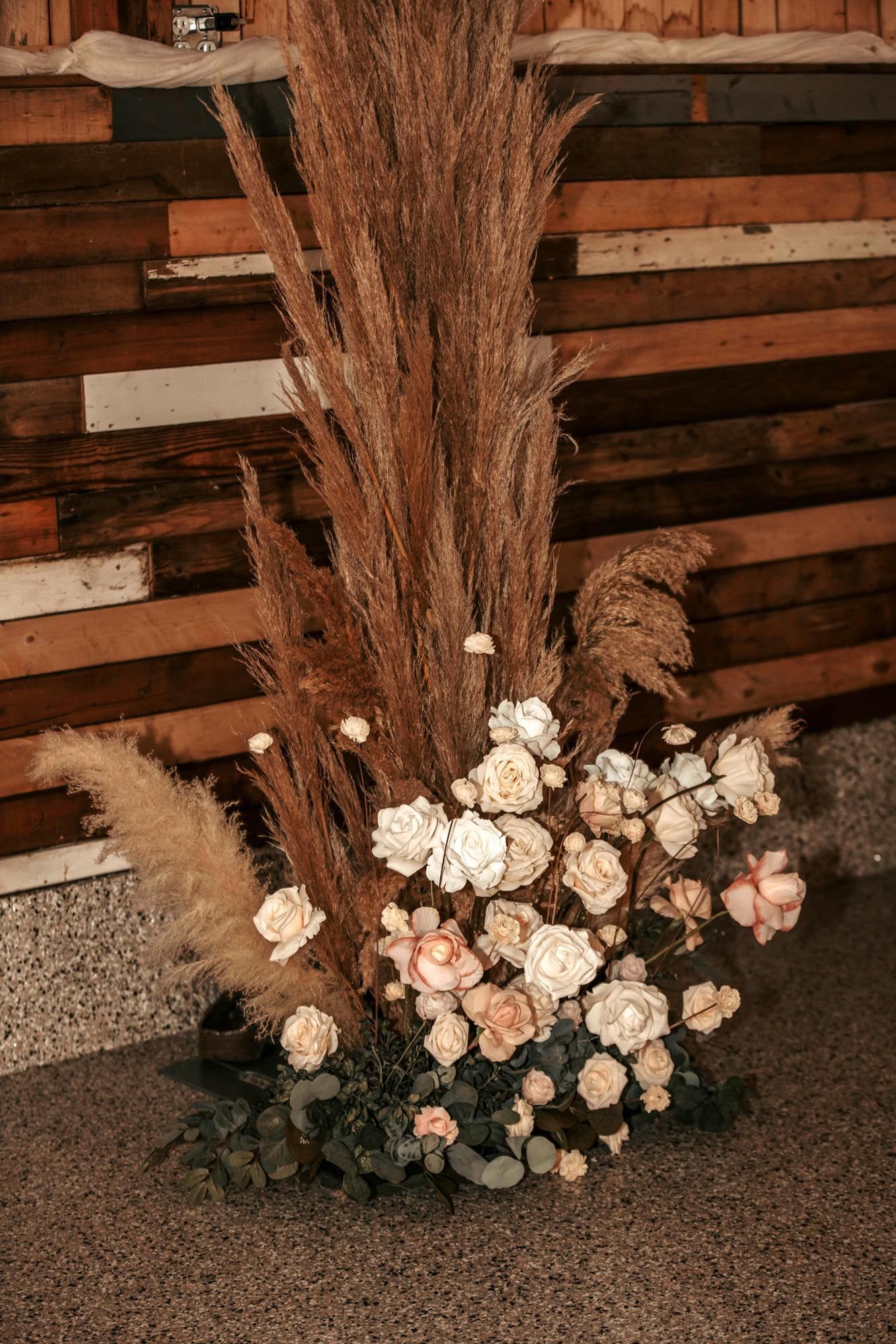 A tall, rustic floral arrangement with dried grasses and beige roses set against a dark wood wall.