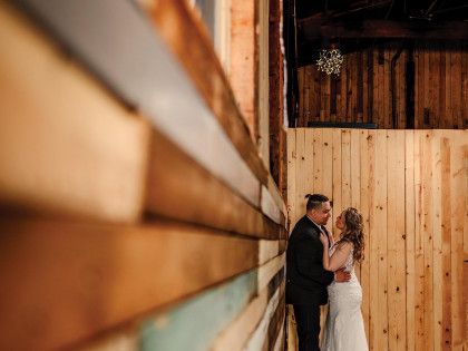 A couple in wedding attire faces each other lovingly against a rustic, vertical wood-plank wall in a dimly lit room.