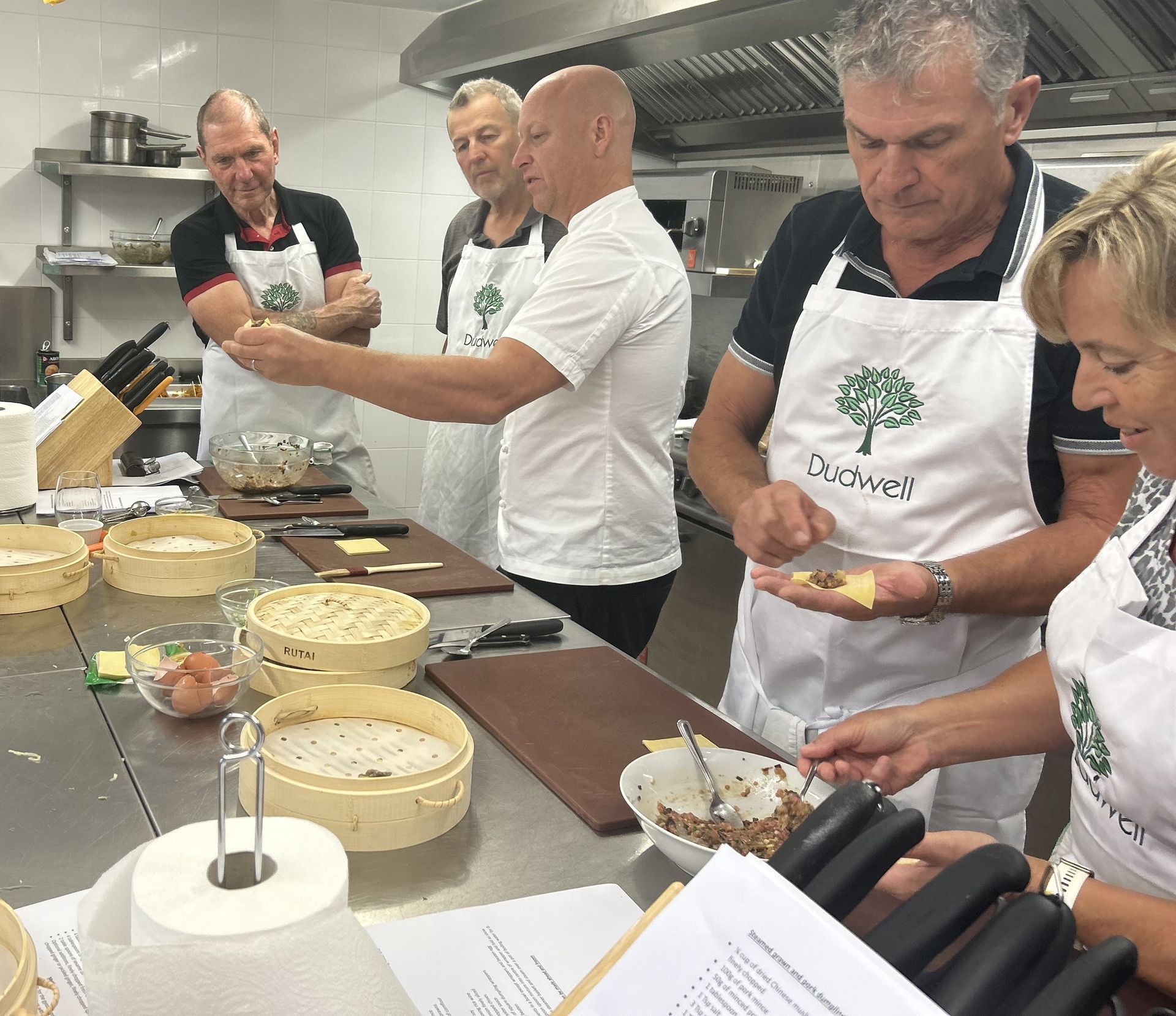 People in a kitchen, learning to cook dumplings. They are wearing aprons and looking at a chef's demonstration.