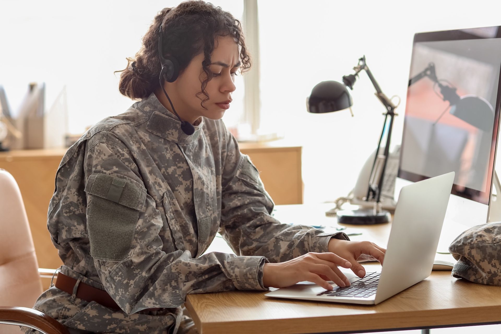 Person typing on a laptop at a desk in a bright office, wearing headphones and a patterned jacket