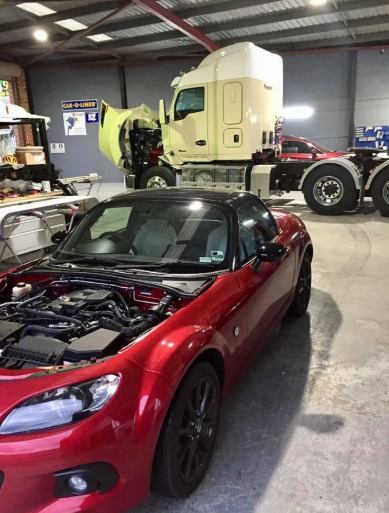 A red car parked in a shed with the bonnet up and trucks in the background — Seabreeze Auto Body in Coffs Harbour, NSW
