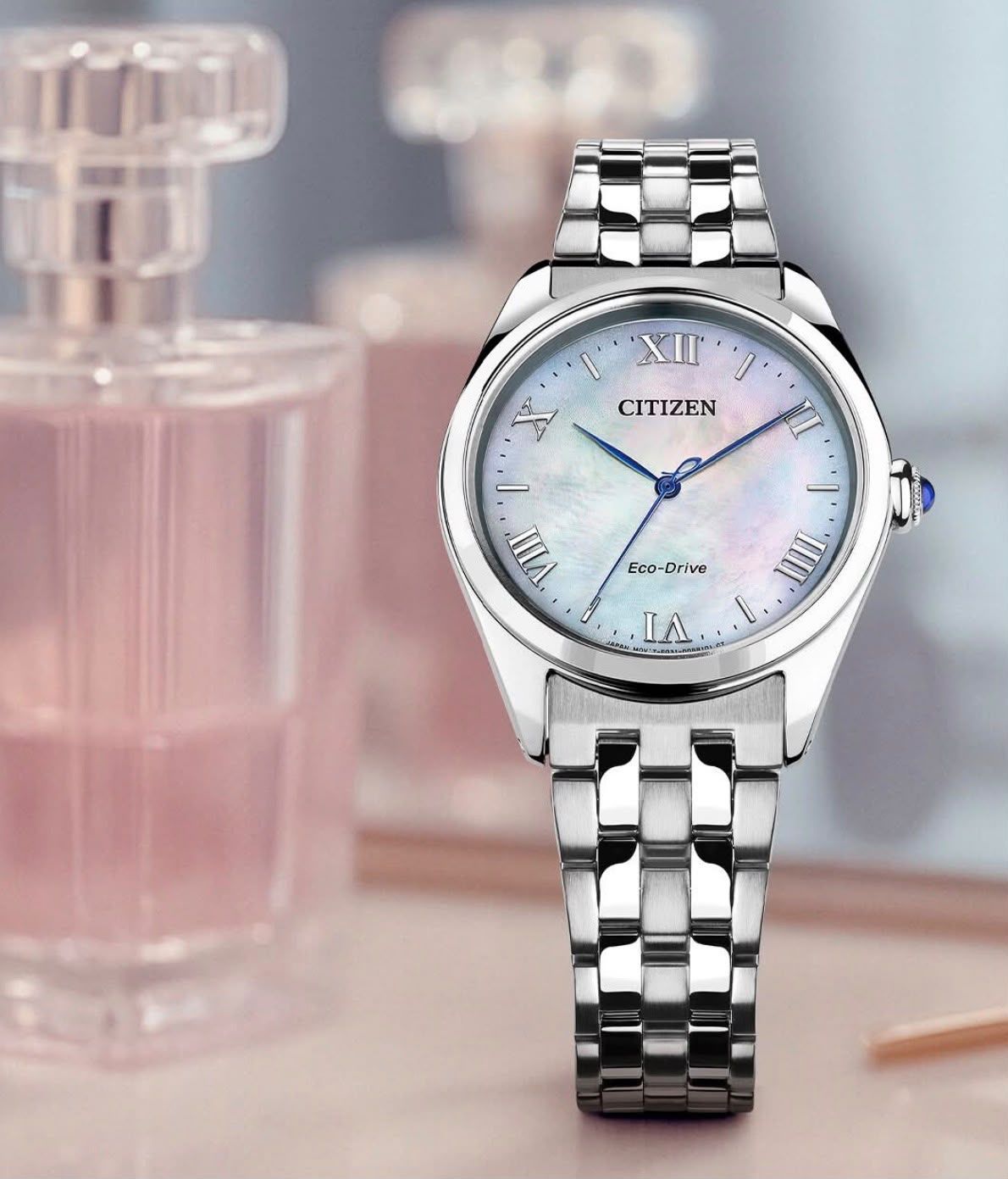 Orologio argentato Citizen con quadrante iridescente accanto a flaconi di profumo rosa