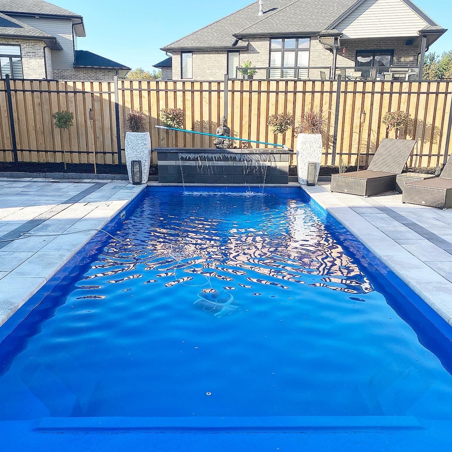 Blue rectangular swimming pool, backyard setting with wooden fence, waterfall feature, and two houses in the background.