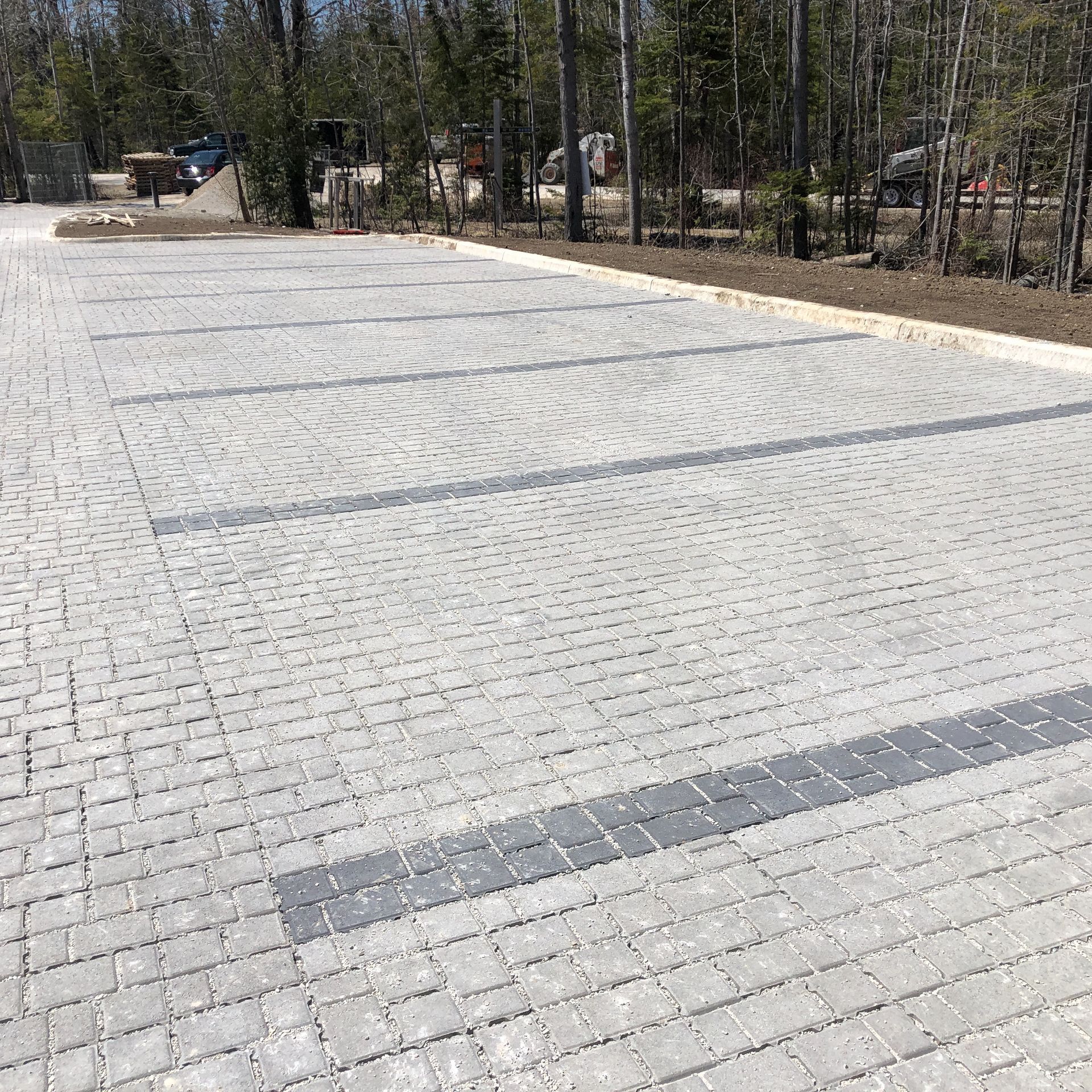 Gray brick parking lot with dark gray bands, trees in the background.