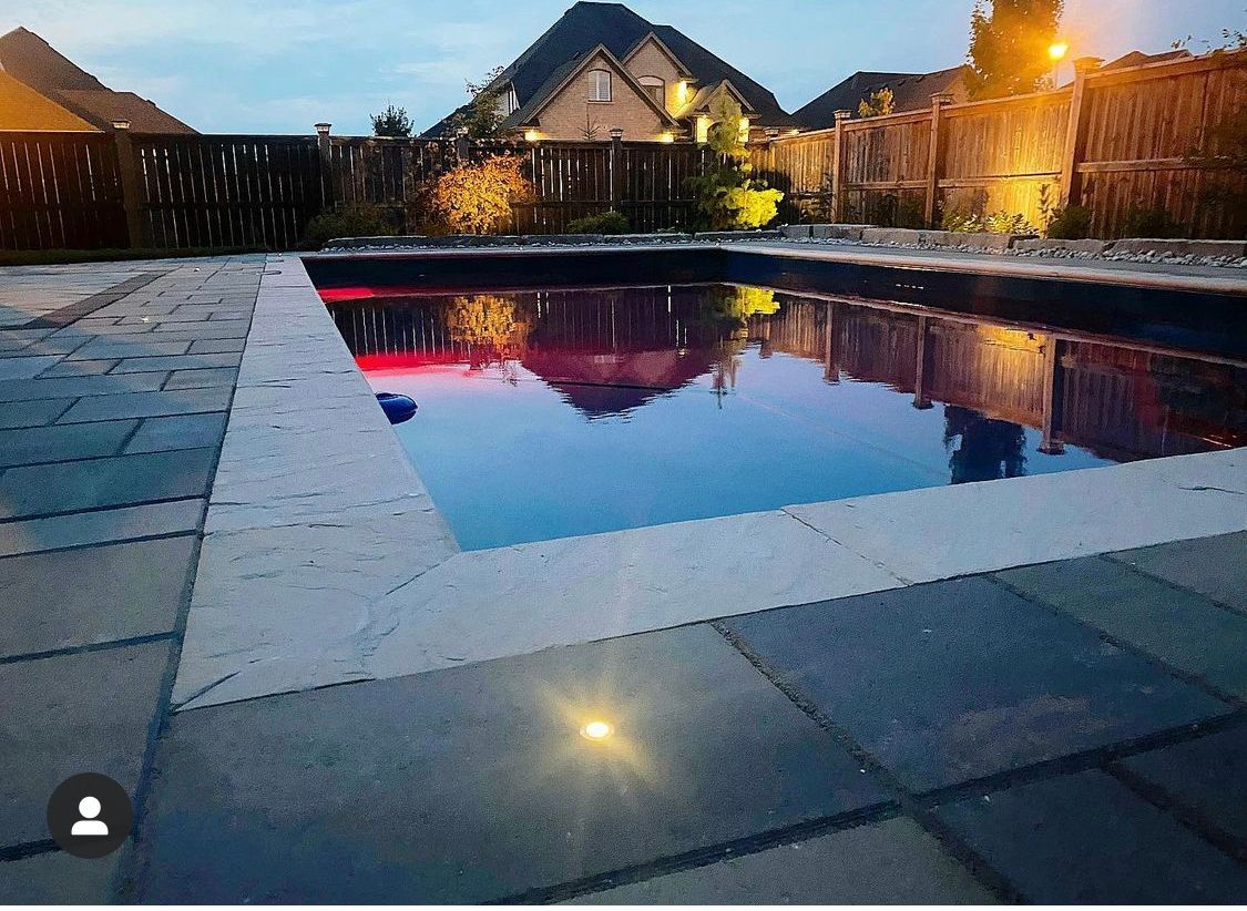 Pool at dusk, reflecting lights and surrounding houses. Blue water, gray tile, and warm ambient lighting.