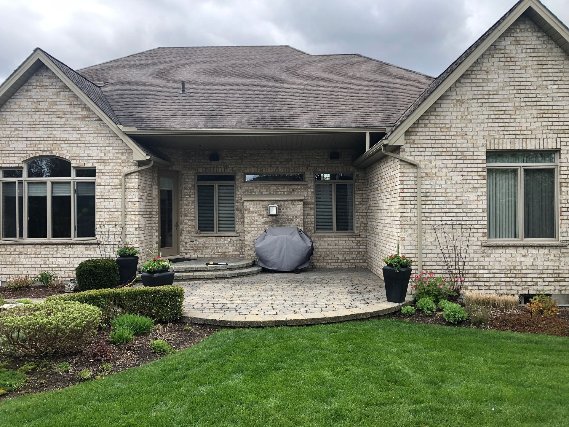 Backyard patio with brick house, stone patio, green grass, and potted plants. Overcast sky.
