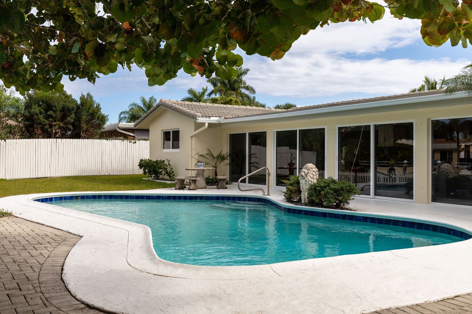 Backyard with pool, beige house, large windows, blue water, green lawn, and trees.