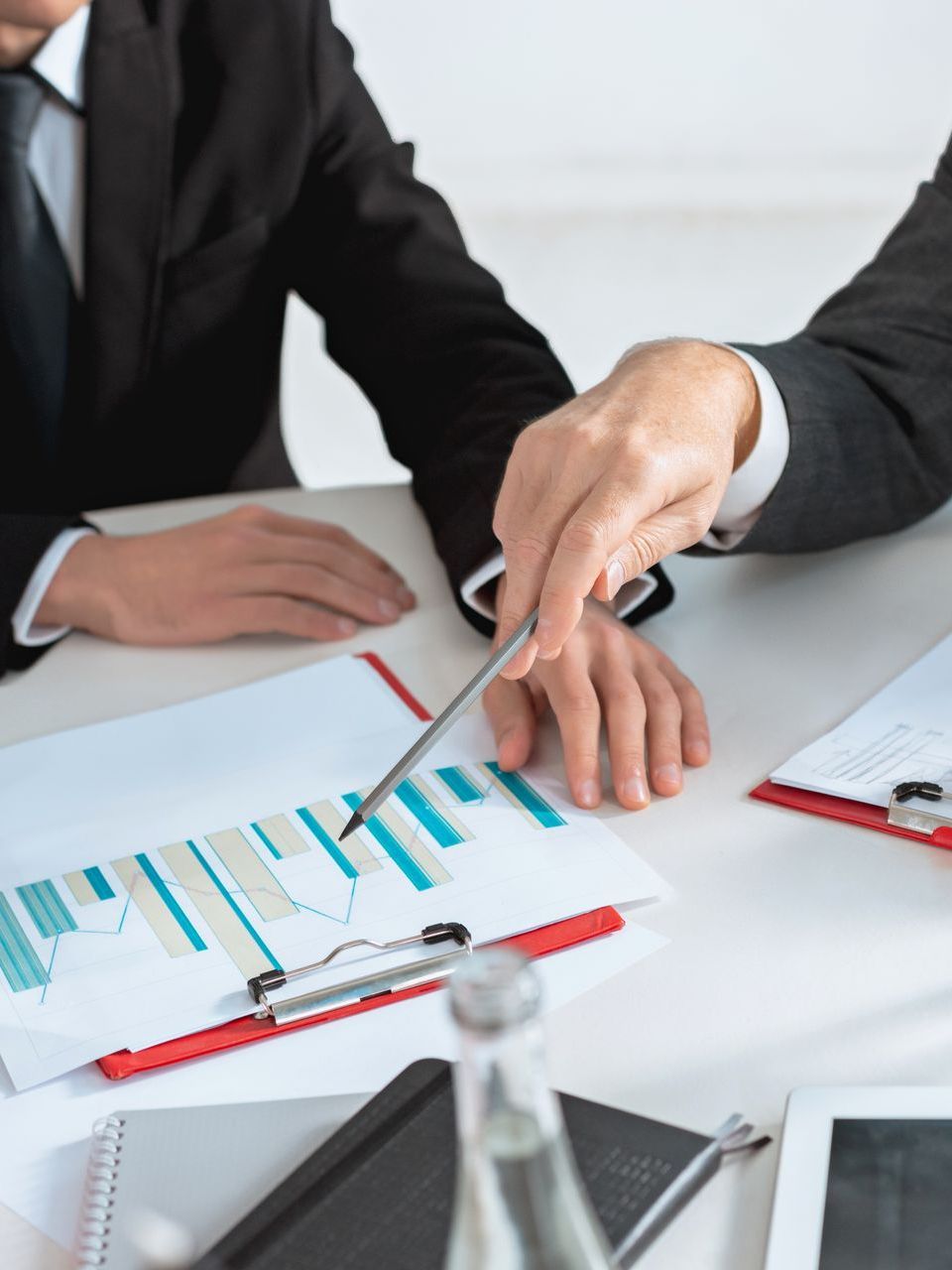 Two businesspeople in suits, one pointing at a bar graph on a paper in a meeting.