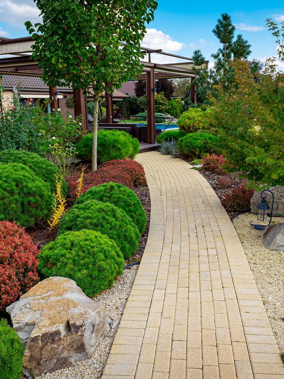 Stone path winds through a colorful garden with green and red shrubs, leading towards a wooden pergola.