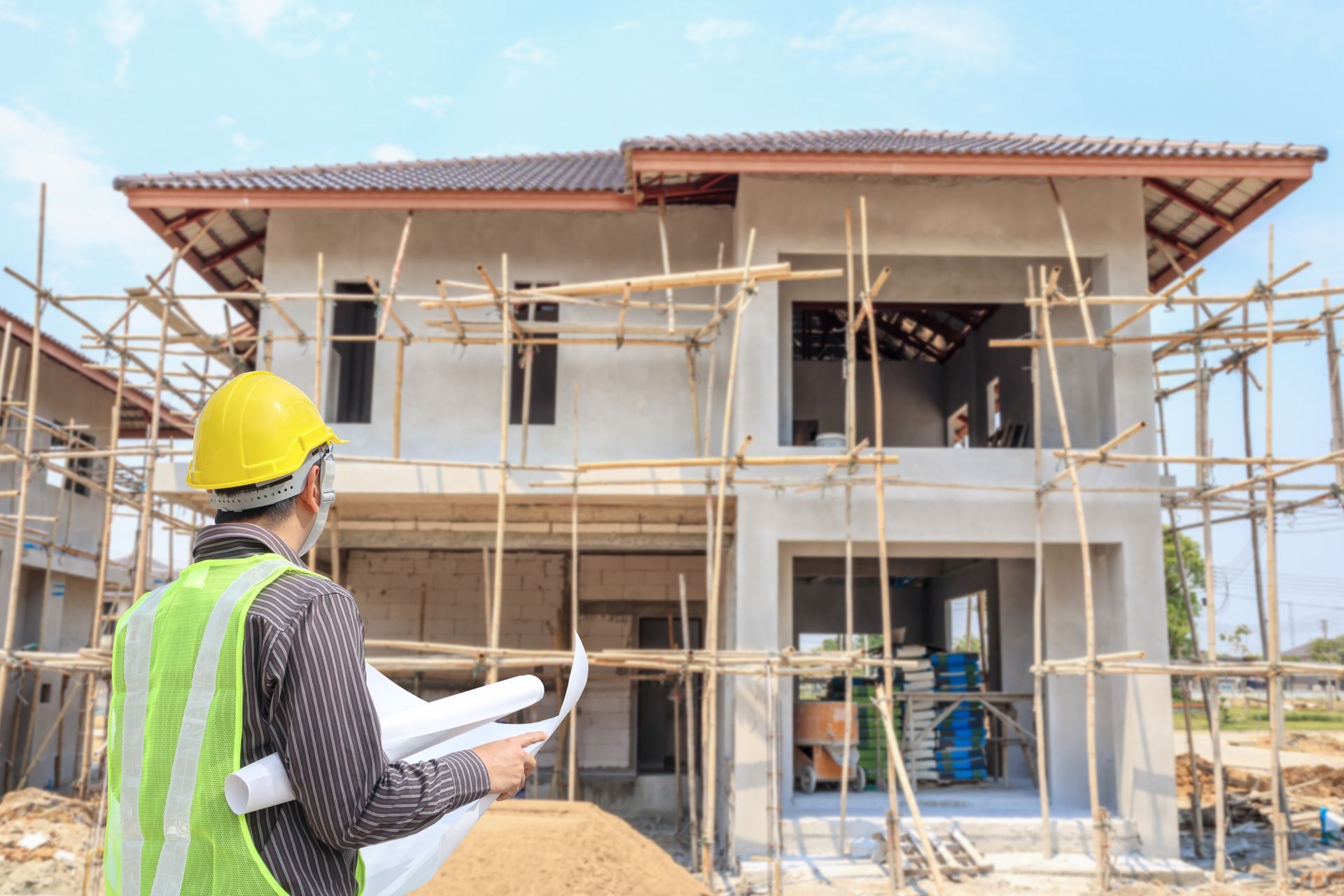 Construction worker inspecting house under construction; wearing hard hat, holding blueprints.