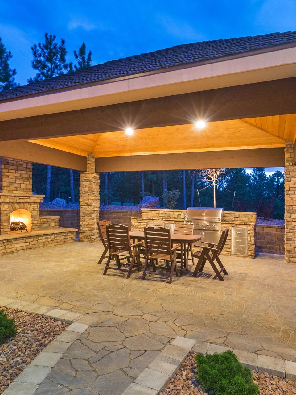 Stone patio with covered seating, fireplace, and outdoor kitchen. Dusk setting, blue sky, and warm lights.