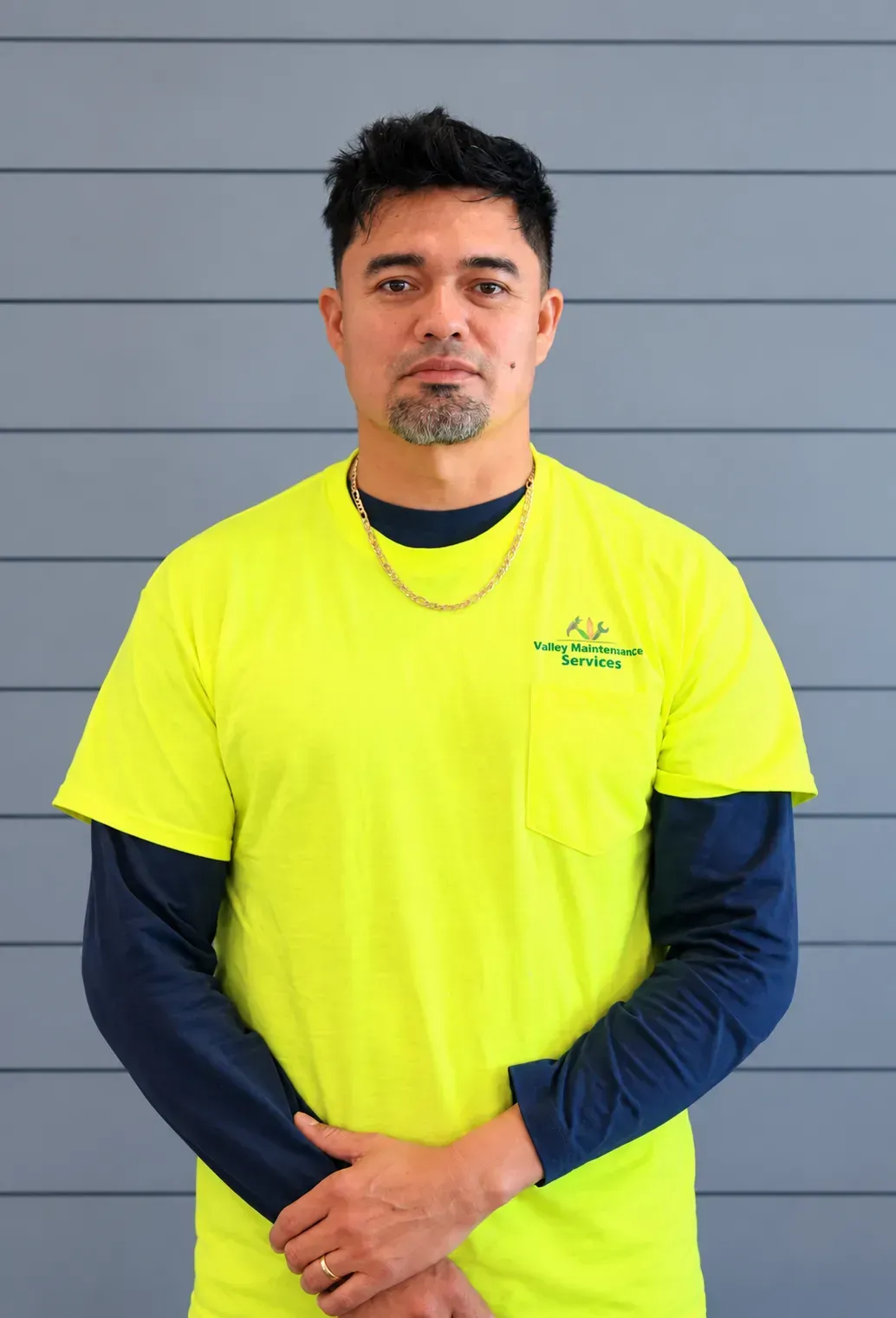 Person in a neon yellow shirt and navy sleeves stands against a gray wall with hands clasped.