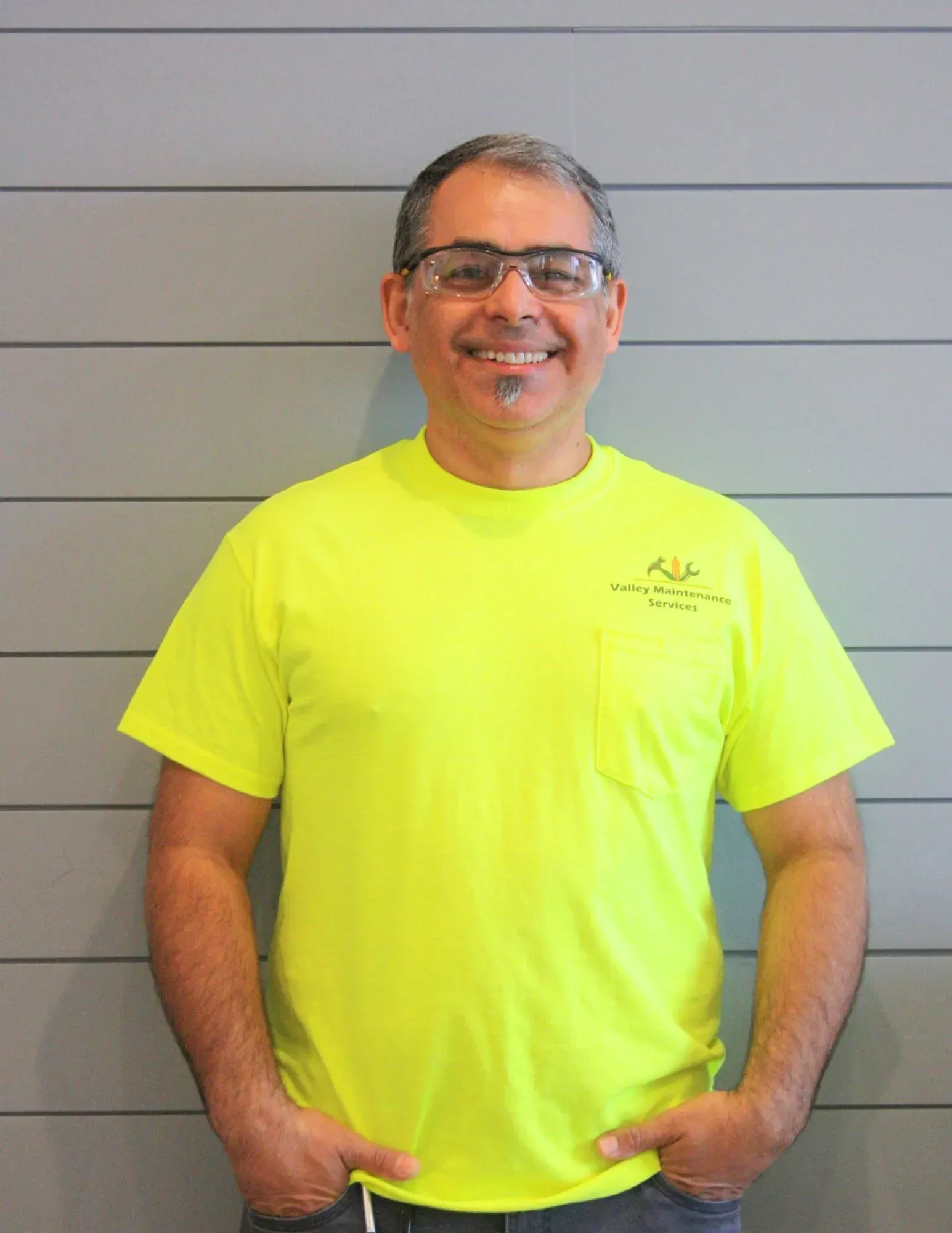 Smiling man in neon yellow T-shirt standing against a gray wall, hands in pockets.