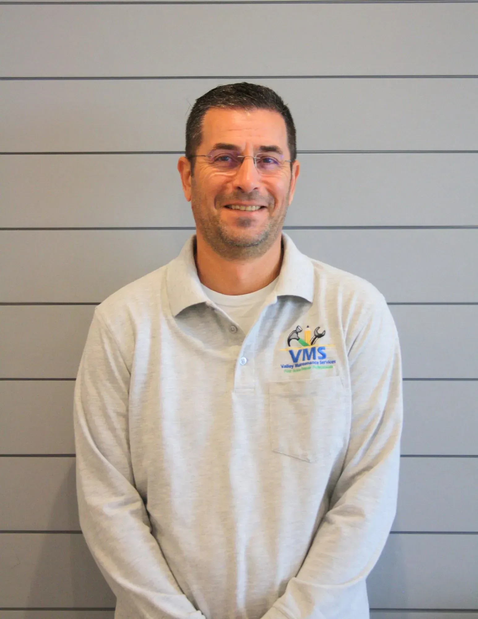 Man standing against a gray wall, wearing a light gray VMS polo shirt and smiling