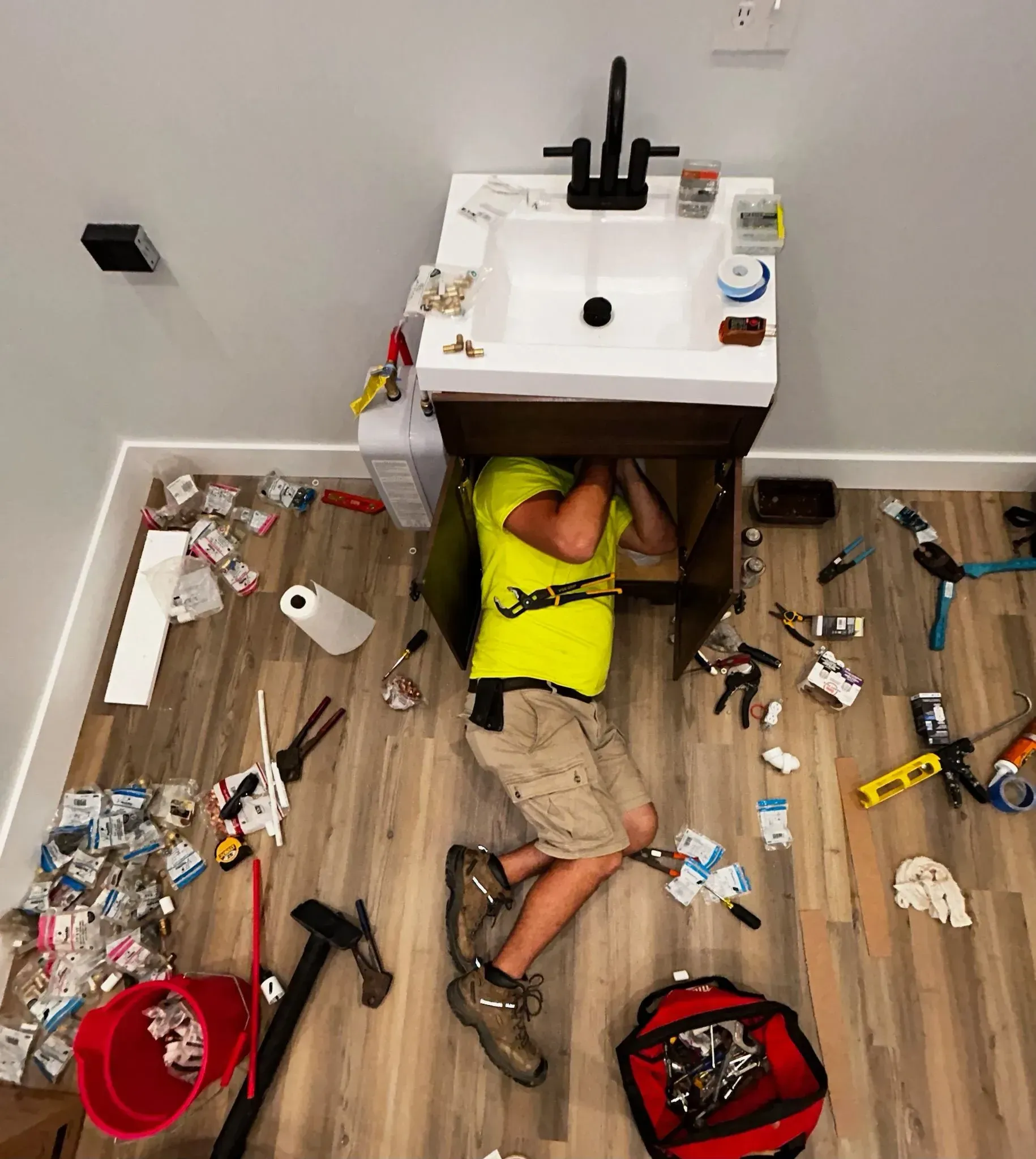 Person lying under a bathroom sink amid tools and debris, wearing a bright yellow shirt and khaki shorts.