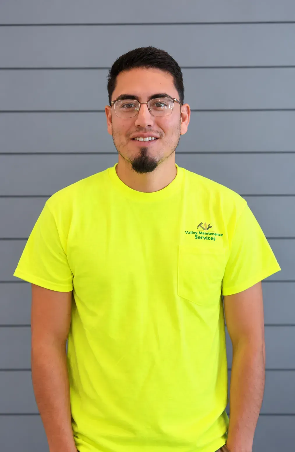Man standing in front of a gray wall, wearing a bright neon yellow T-shirt and glasses.