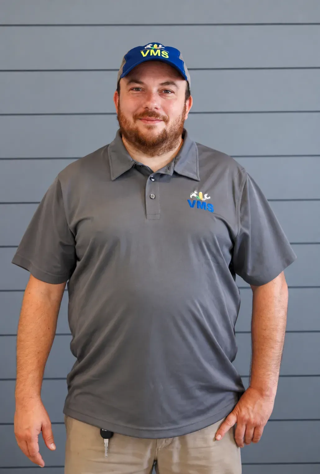 Man in gray polo and blue cap standing against a gray wall, wearing a company logo.